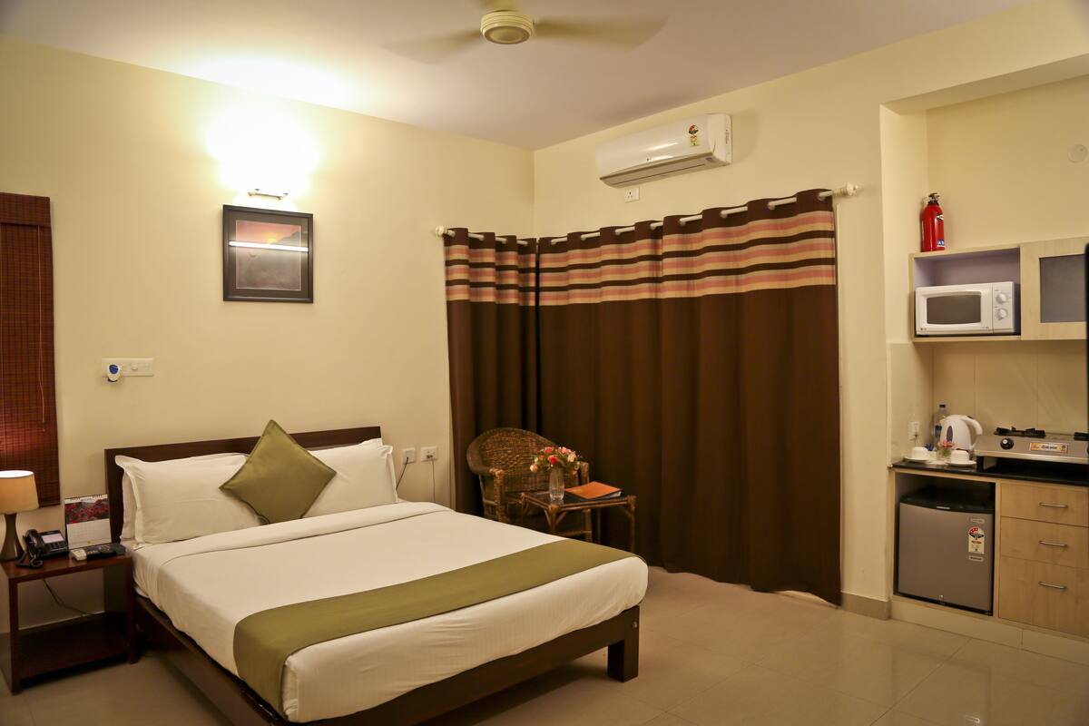 A hotel room with a bed, a nightstand, a chair, a small table, a microwave, and a refrigerator.