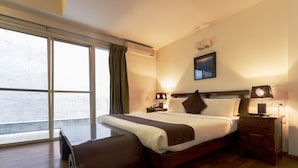 A hotel room with a large window, a bed with a wooden headboard, a bedside table with a lamp, and a bench. - Maple Suites Serviced Apartments (Bengaluru)