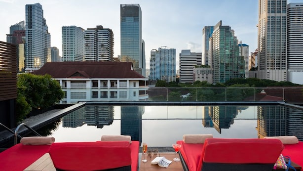 Una piscina en la azotea con sillas reclinables rojas y una mesa, con vistas a un paisaje urbano con edificios modernos y altos.