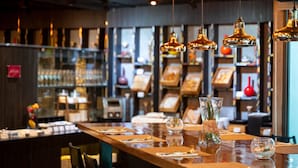 Interior de un restaurante con una larga mesa de madera dispuesta para comer, luces colgantes y estantes con diversos objetos al fondo.
