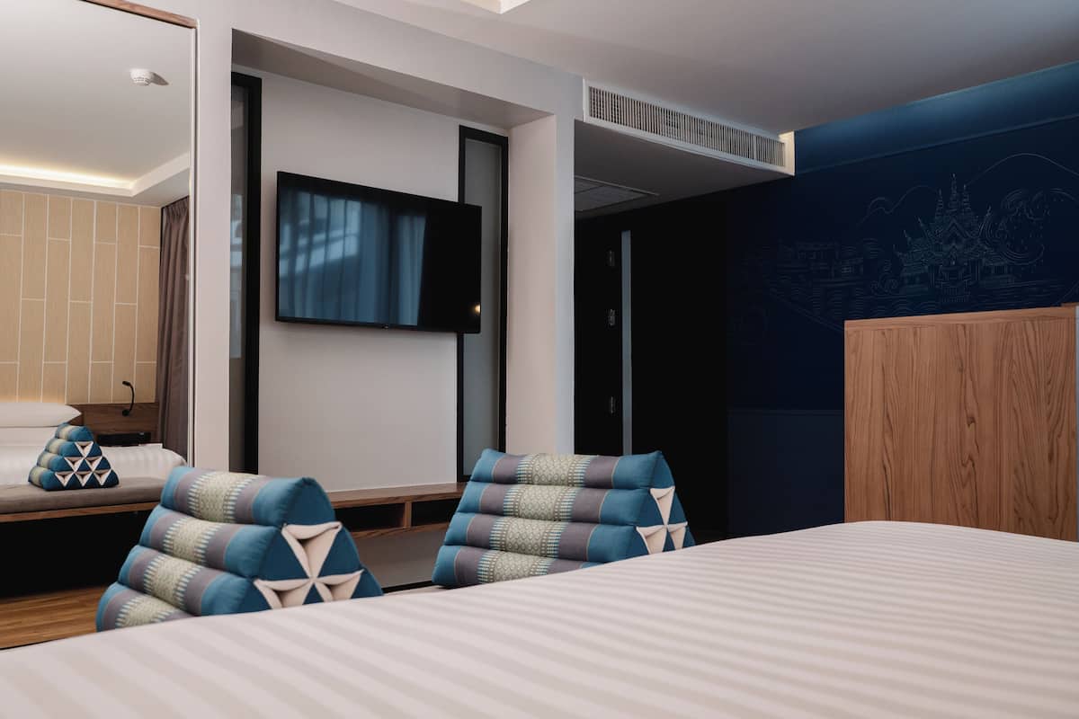 A modern hotel room with a large bed, two armchairs, a flat-screen TV mounted on the wall, and a wooden wardrobe.
