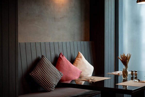Un interior moderno con un panel de madera oscura, un banco acolchado con cojines estampados, una mesa de vidrio con un florero y saleros, y una ventana con luz natural.