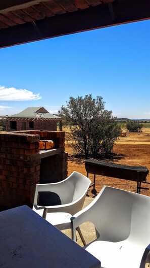 A patio area with a brick barbecue grill, white plastic chairs, and a view of a brick building and trees. - Sangiro Game Lodge (Bloemfontein)