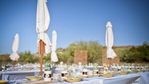 Montaje de comedor frente a la playa, con mesas cubiertas de manteles blancos, cada una con una pequeña maceta y un vaso de agua.