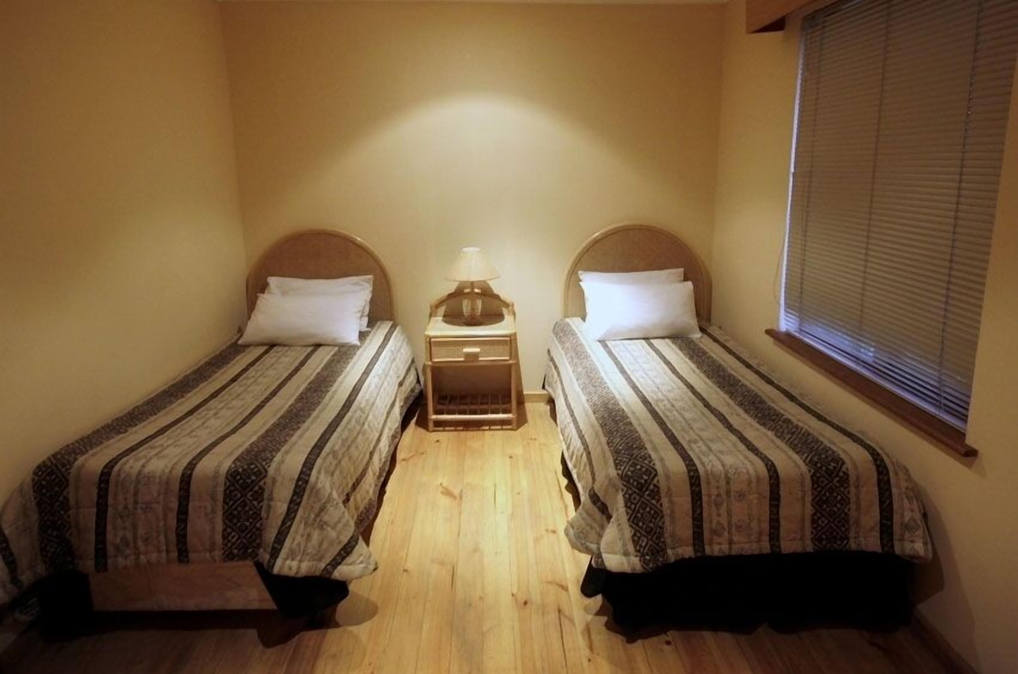 Une petite chambre avec deux lits simples, une table de chevet avec une lampe et une fenêtre avec des stores.