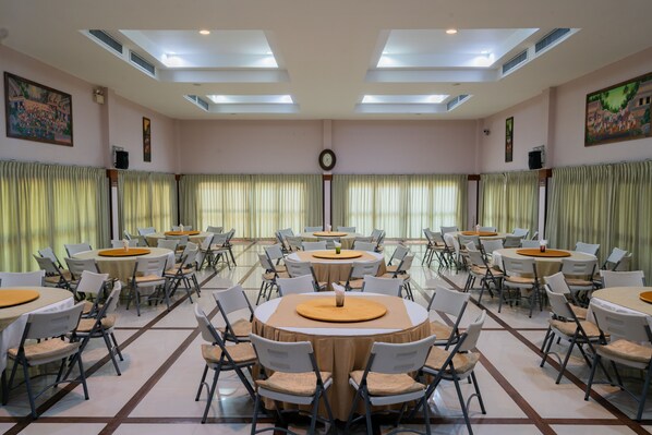 A spacious dining hall with multiple round tables and chairs, large windows with curtains, and a clock on the wall. - Maikaew Damnoen Resort (Damnoen Saduak)