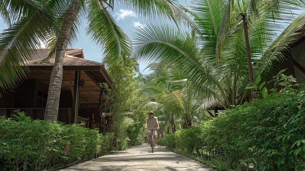 A pathway flanked by lush greenery and a traditional building with a thatched roof. - Maikaew Damnoen Resort (Damnoen Saduak)