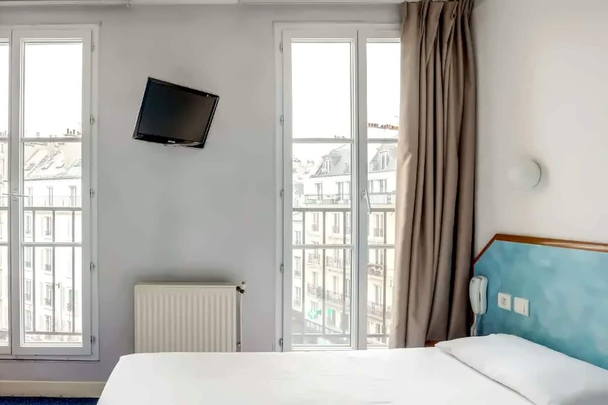 Habitación de hotel con cama, televisor montado en la pared, radiador, teléfono y ventanas amplias con cortinas.