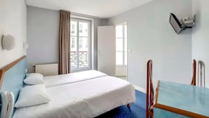 A hotel room with a bed, a table, a chair, a window with curtains, and a wall-mounted device.