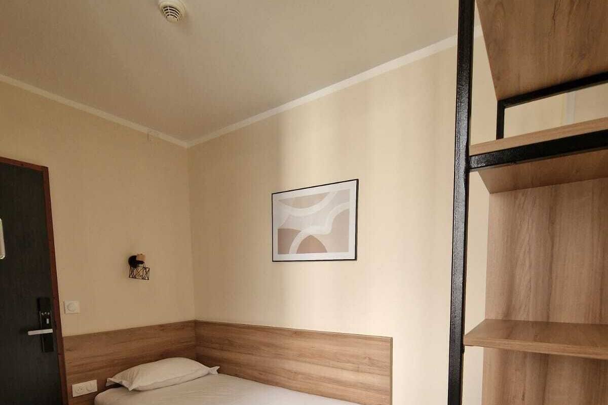 Une chambre à coucher avec un lit en bois, une petite table de chevet, une étagère fixée au mur et une porte.