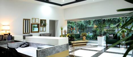 A modern reception area with a marble reception desk, framed certificates on the wall, and a view of a lush garden through large windows.