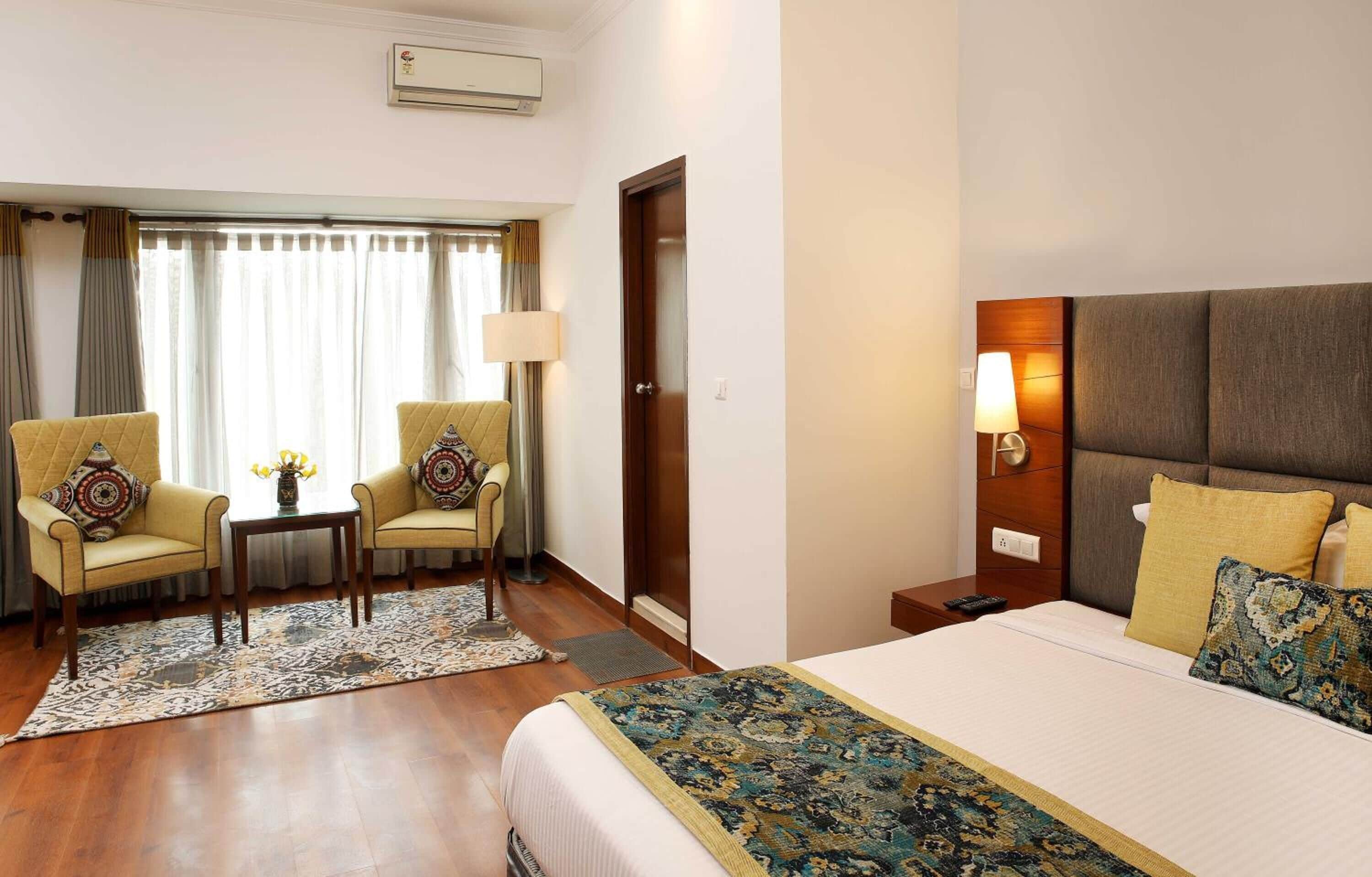 A hotel room with a large bed, two armchairs, a small table with a vase, a nightstand with a lamp, and a door.