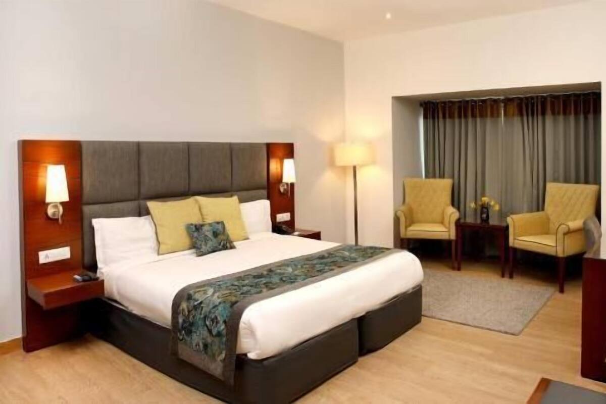 A hotel room with a large bed, bedside tables, a lamp, two armchairs, and a view of the outside.