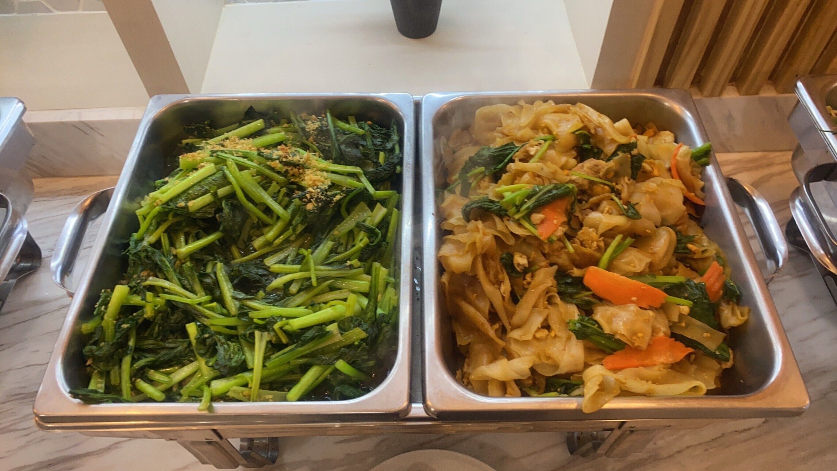 Une table de buffet avec deux plateaux de service en métal, l’un contenant des légumes verts et l’autre des nouilles aux légumes.