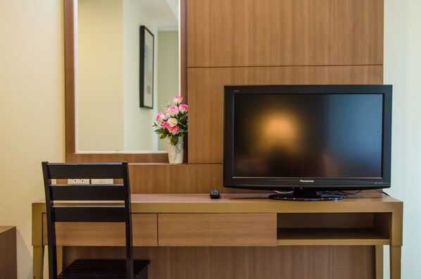 A hotel room with a wooden desk, a black chair, a television, and a vase with flowers.