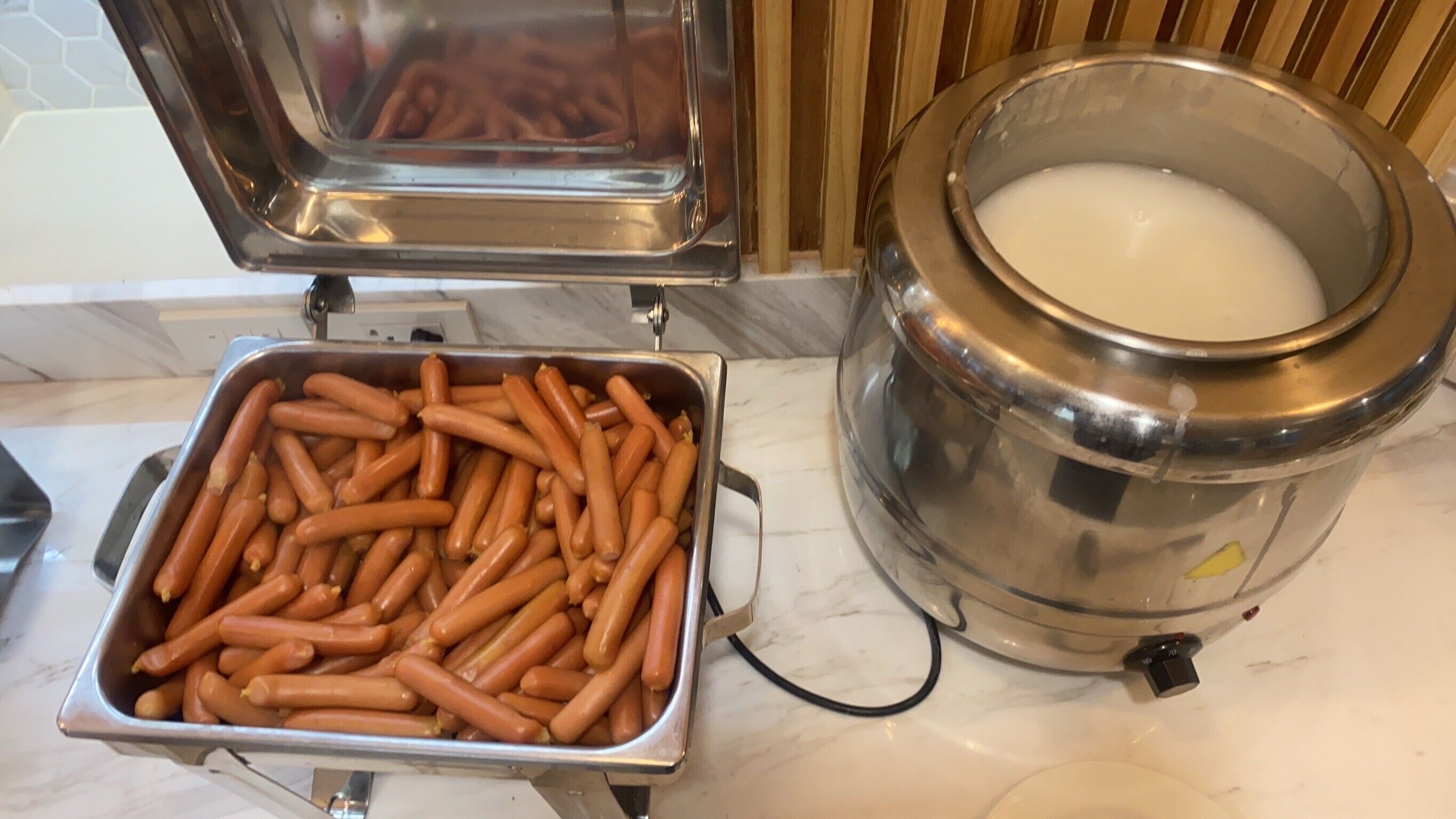 A stainless steel container filled with hot dogs and a milk dispenser on a marble countertop.