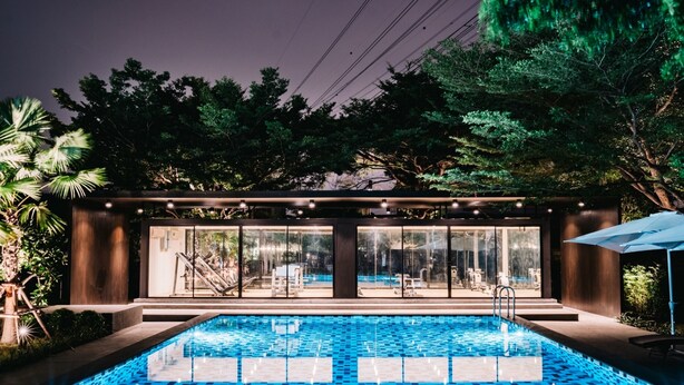 A modern outdoor pool area with a glass-enclosed lounge, surrounded by lush greenery and trees. - Sunday Platinum Suites (Bangkok)