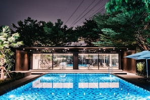 A modern outdoor pool area with a glass-enclosed lounge, surrounded by lush greenery and trees. - Sunday Platinum Suites (Bangkok)