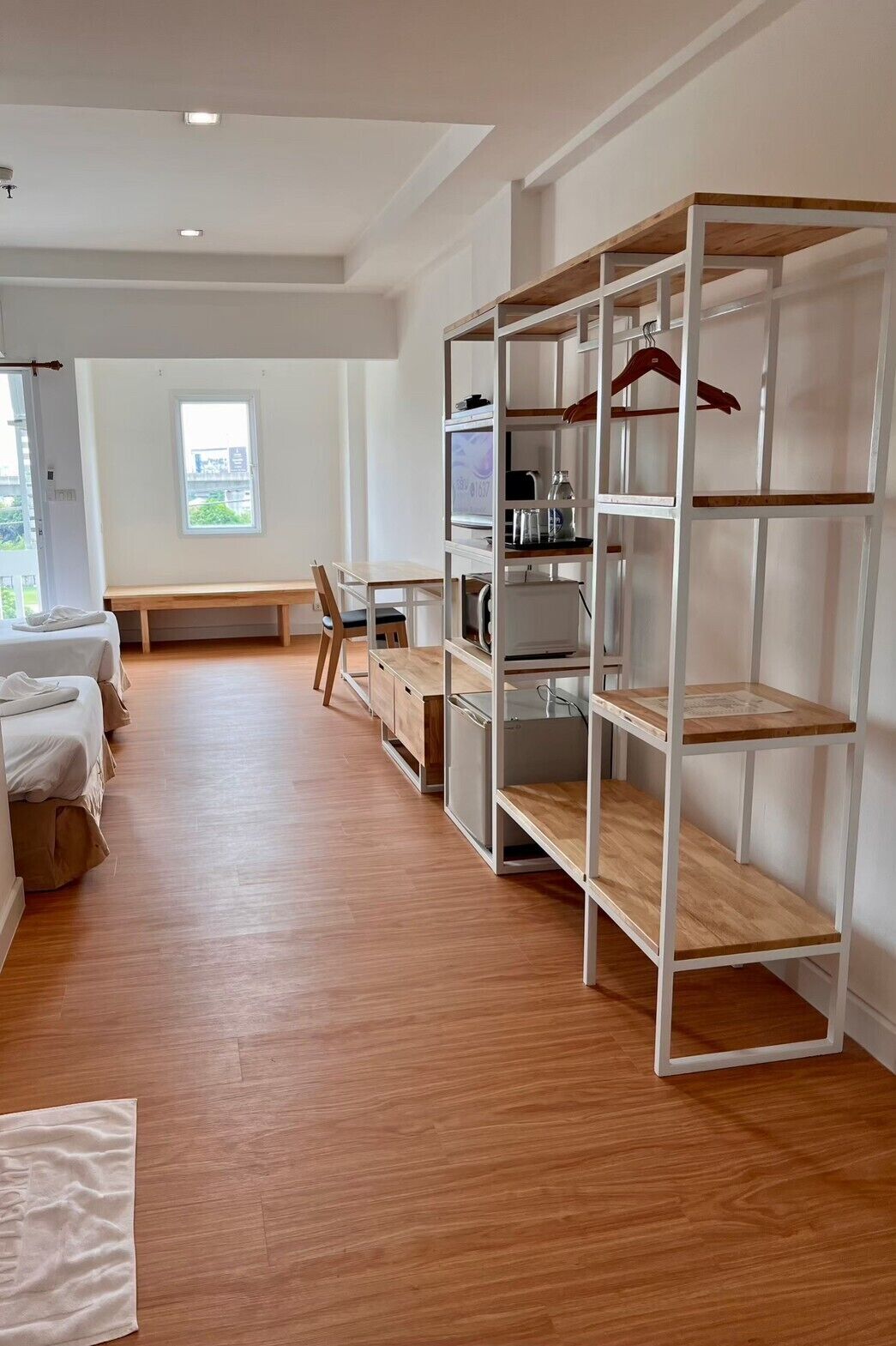 A modern hotel room with a wooden floor, a large window with a city view, a bench, a desk, and a white wardrobe with shelves.