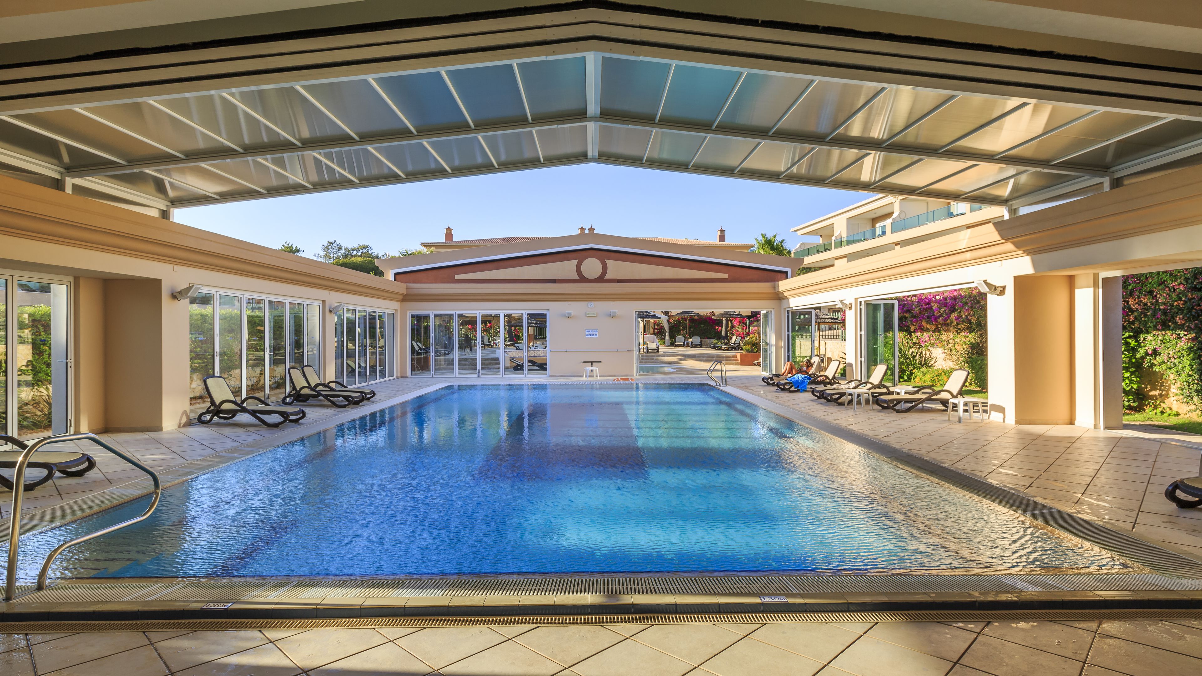 A large indoor swimming pool with clear blue water, surrounded by sun loungers and a covered area with a glass roof.
