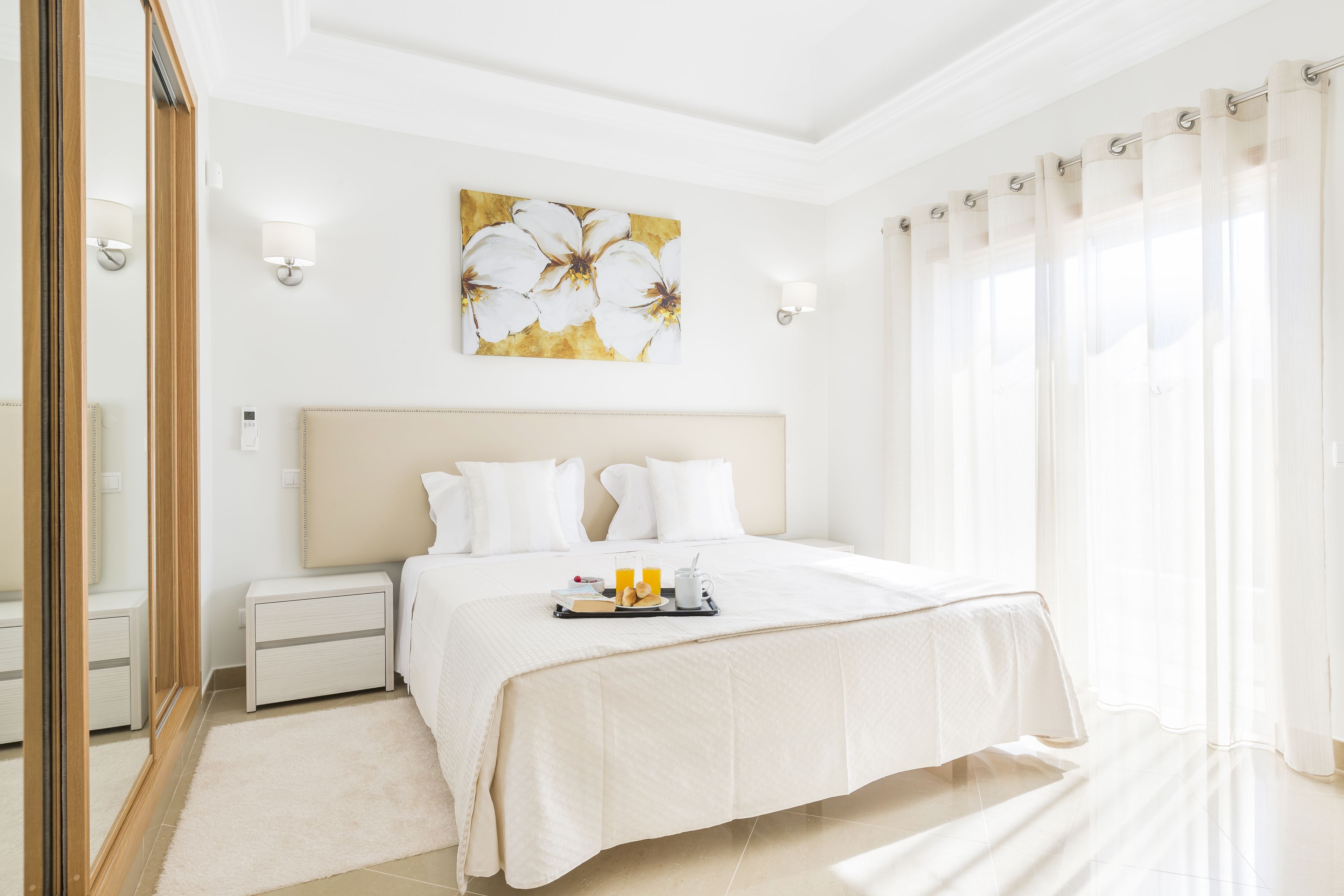 A hotel room with a large bed, a bedside table, a nightstand, a mirror, and a window with curtains.