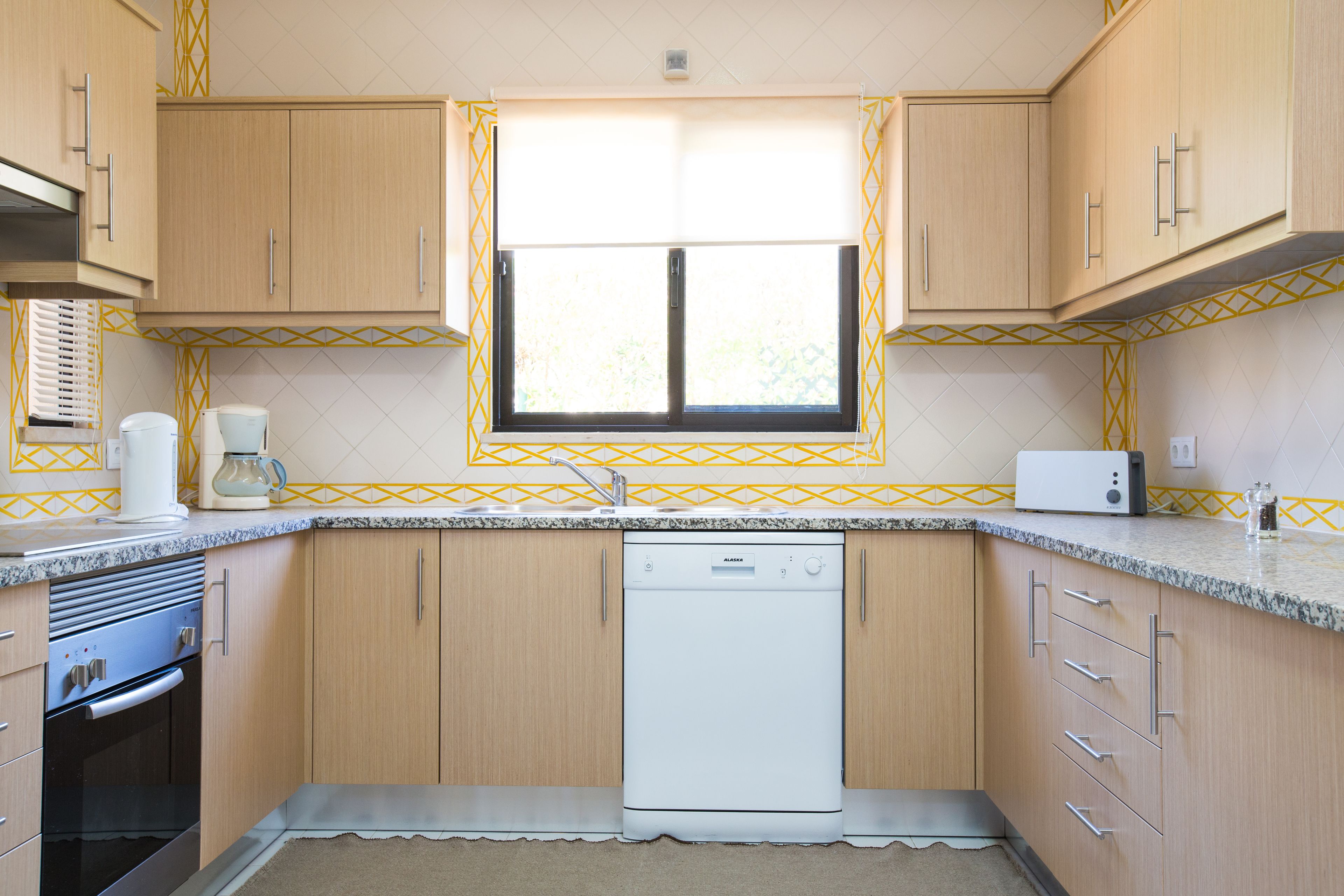 A compact kitchen with wooden cabinets, a built-in dishwasher, and a window with a yellow patterned curtain.