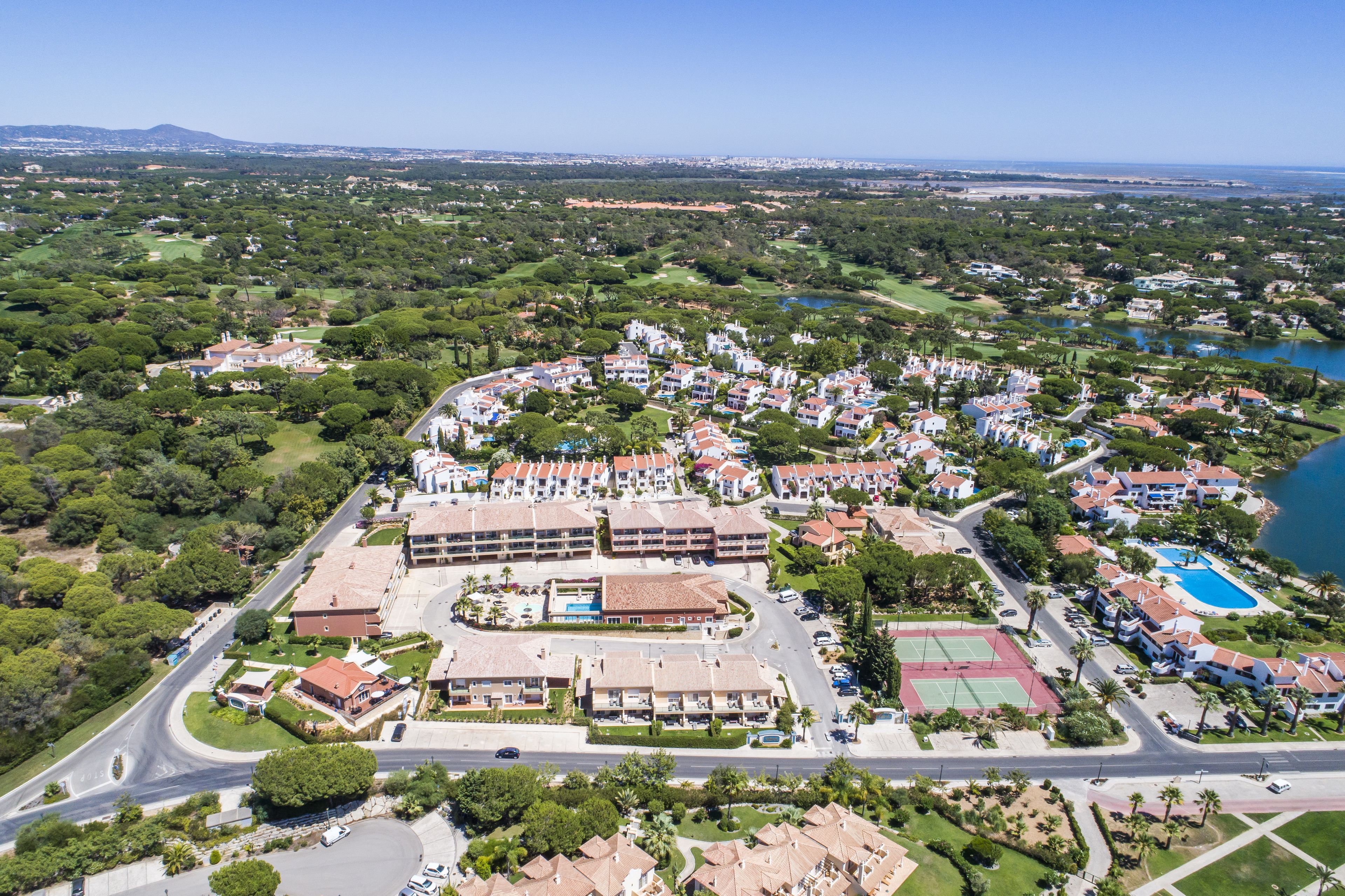 An aerial view of a resort with multiple buildings, a large pool, tennis courts, and a surrounding landscape with trees and a lake.