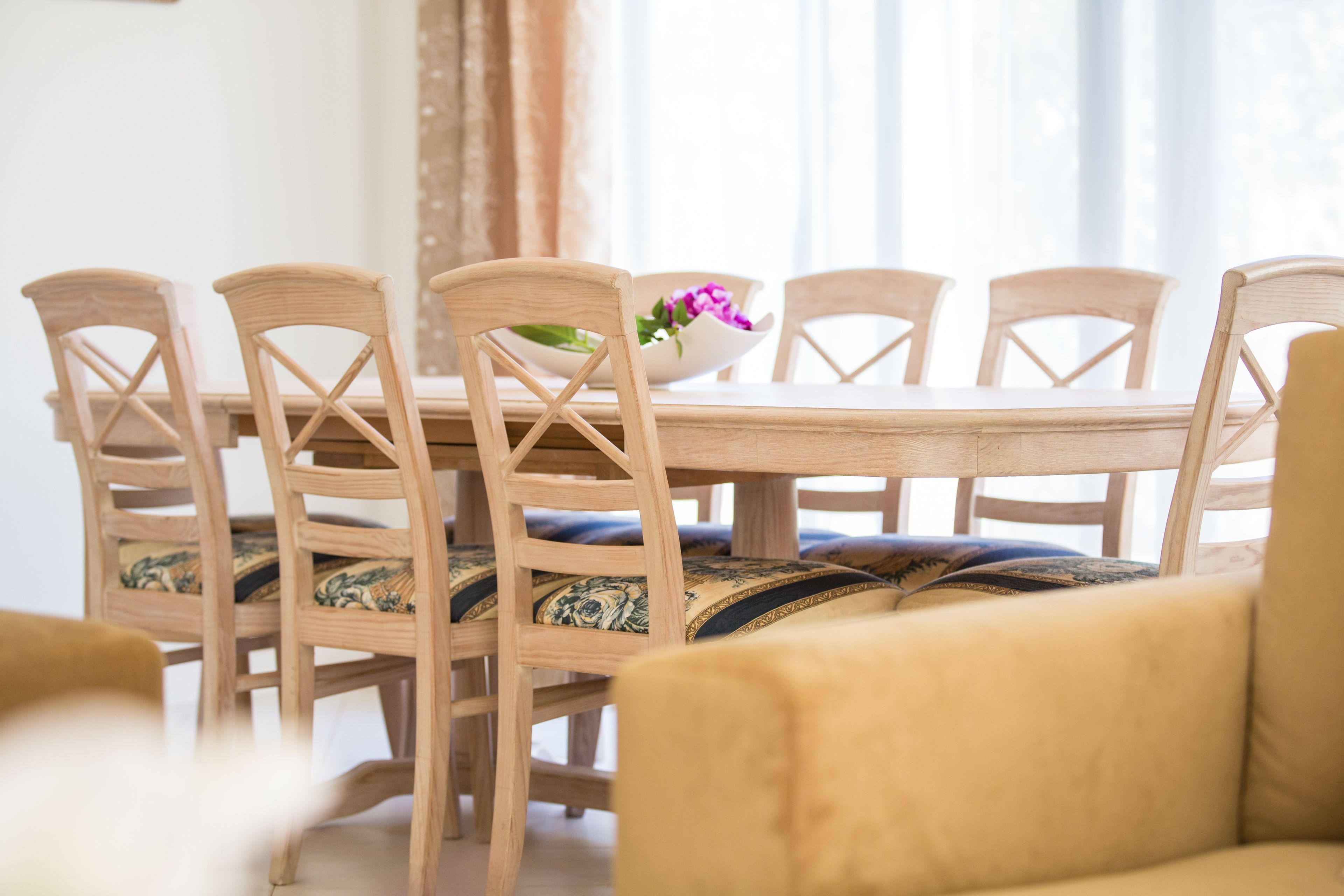 A dining table with six wooden chairs, a bowl with flowers, and a sofa in the foreground.