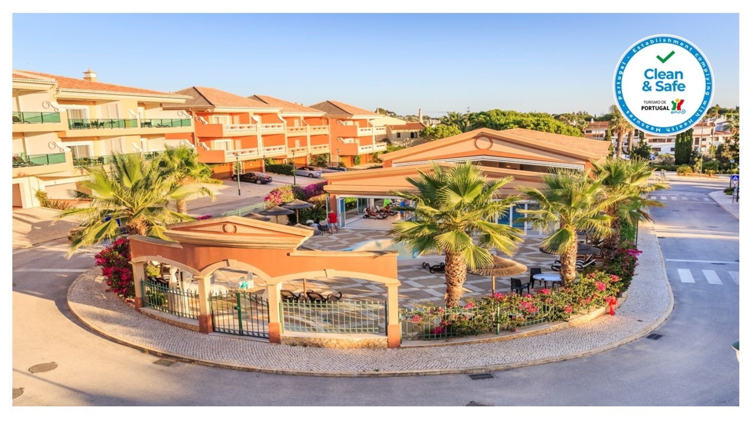 A hotel with multiple buildings, a covered outdoor area with palm trees, and a clear sky.
