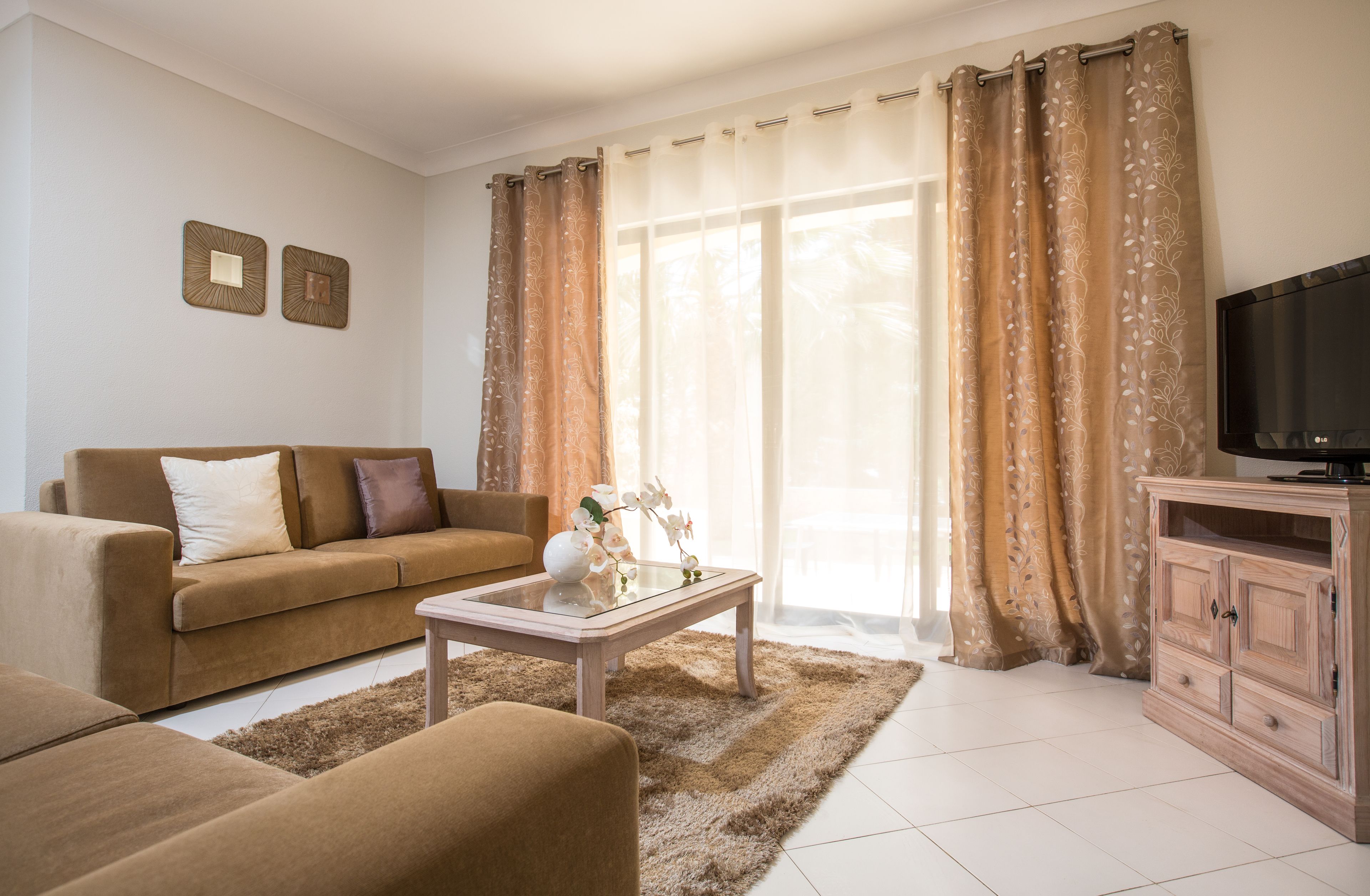 A living room with a sofa, coffee table, and TV stand, featuring a large window with curtains.