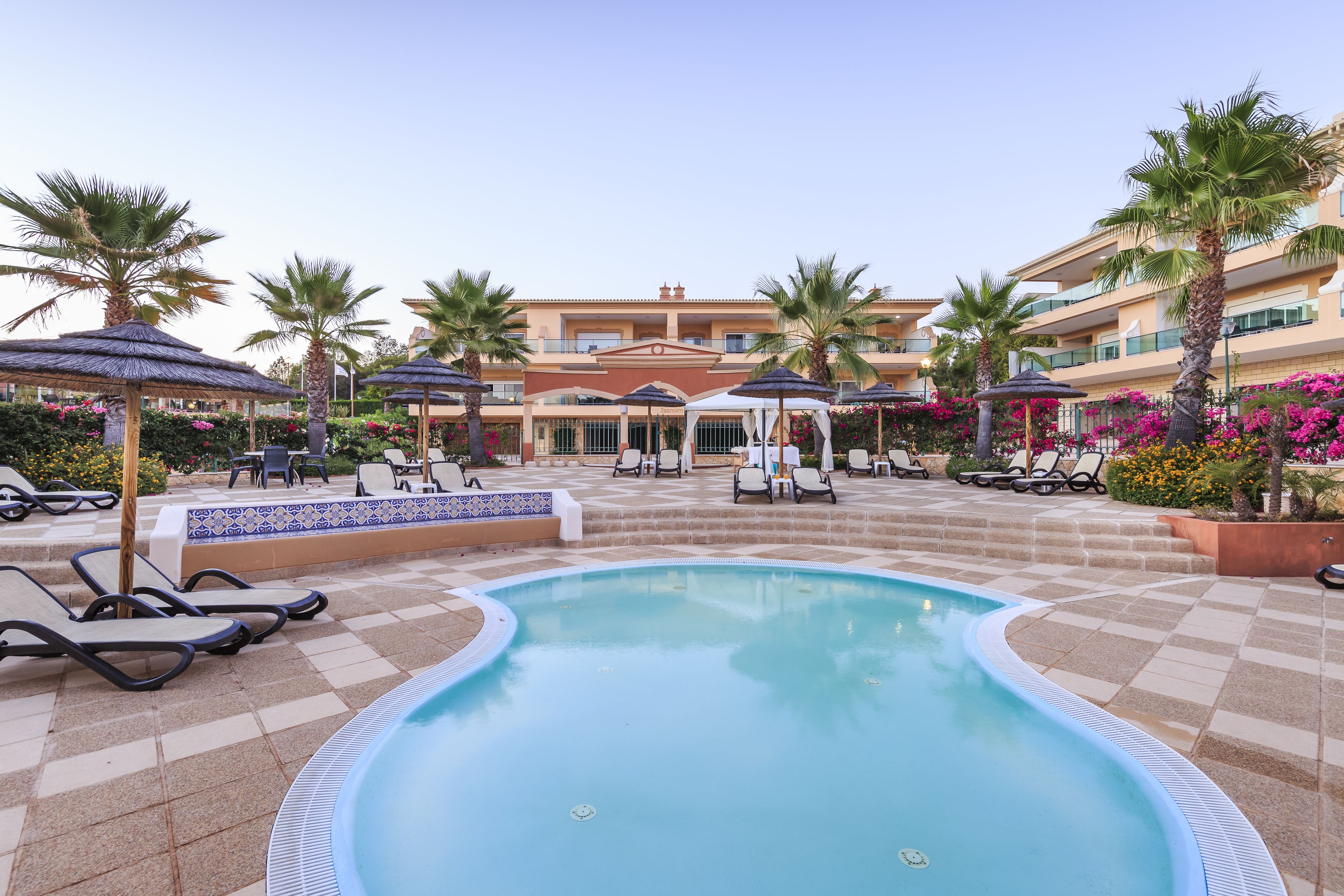 A pool area with lounge chairs and umbrellas, a building with balconies, and a clear sky.