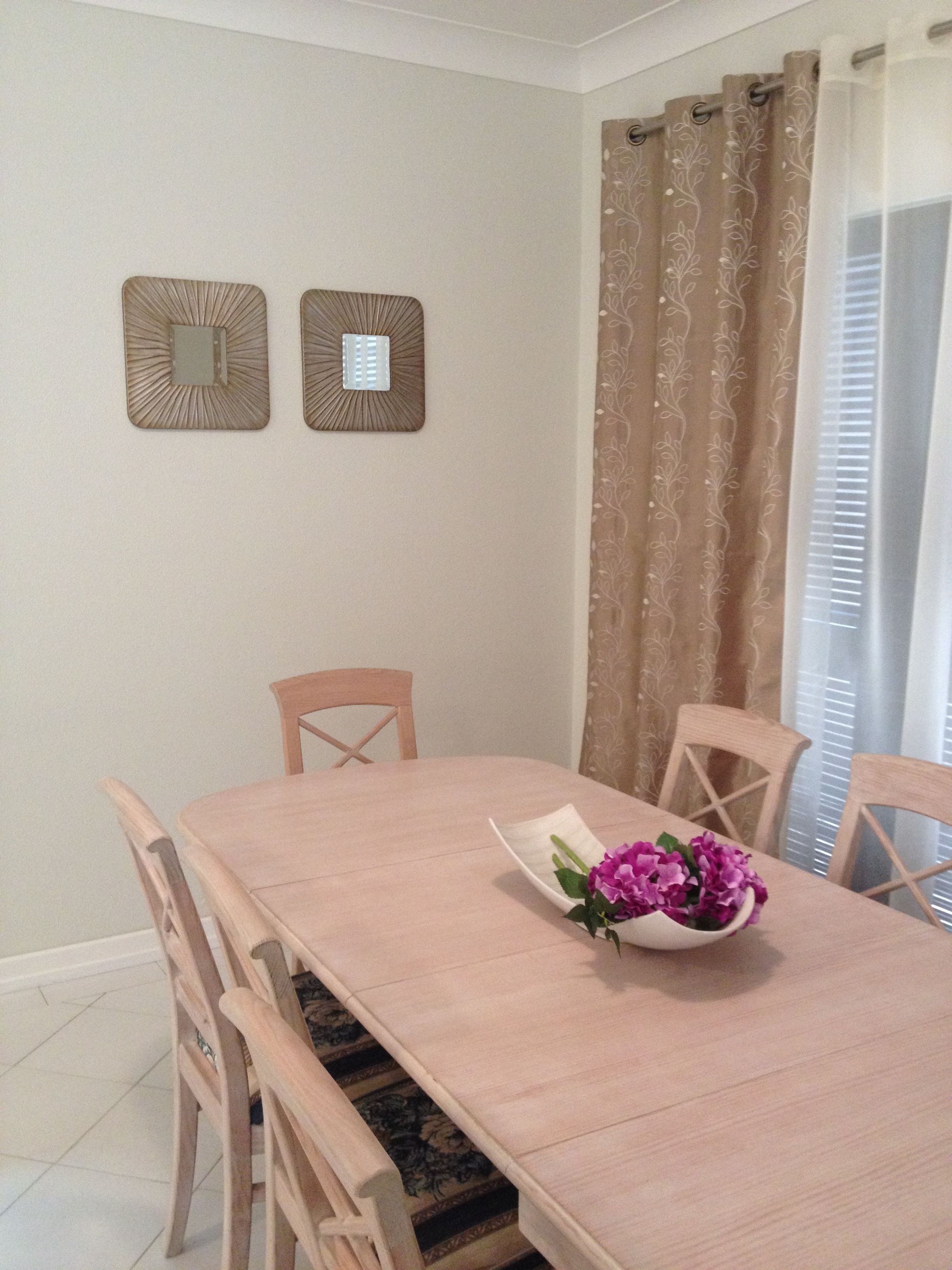Un comedor con una mesa rectangular, cuatro sillas de madera, dos espejos decorativos colgantes y una ventana con cortinas.