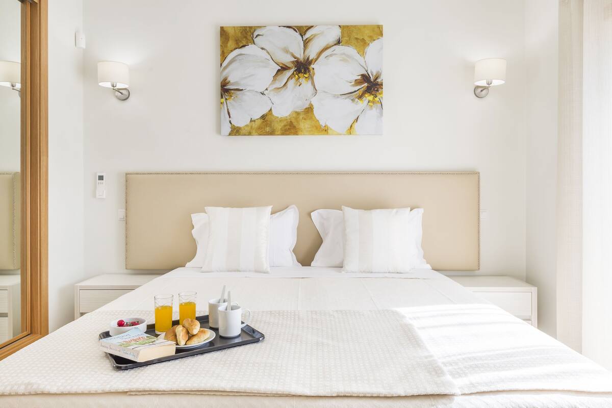 Une chambre d’hôtel avec un grand lit, du linge de lit blanc, un plateau avec des articles pour le petit-déjeuner et un tableau représentant des fleurs accroché au mur.