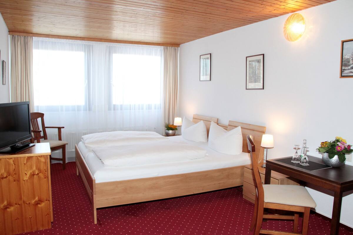 Quarto de hotel com uma cama grande, uma escrivaninha, uma cadeira, uma televisão e uma janela com cortinas.