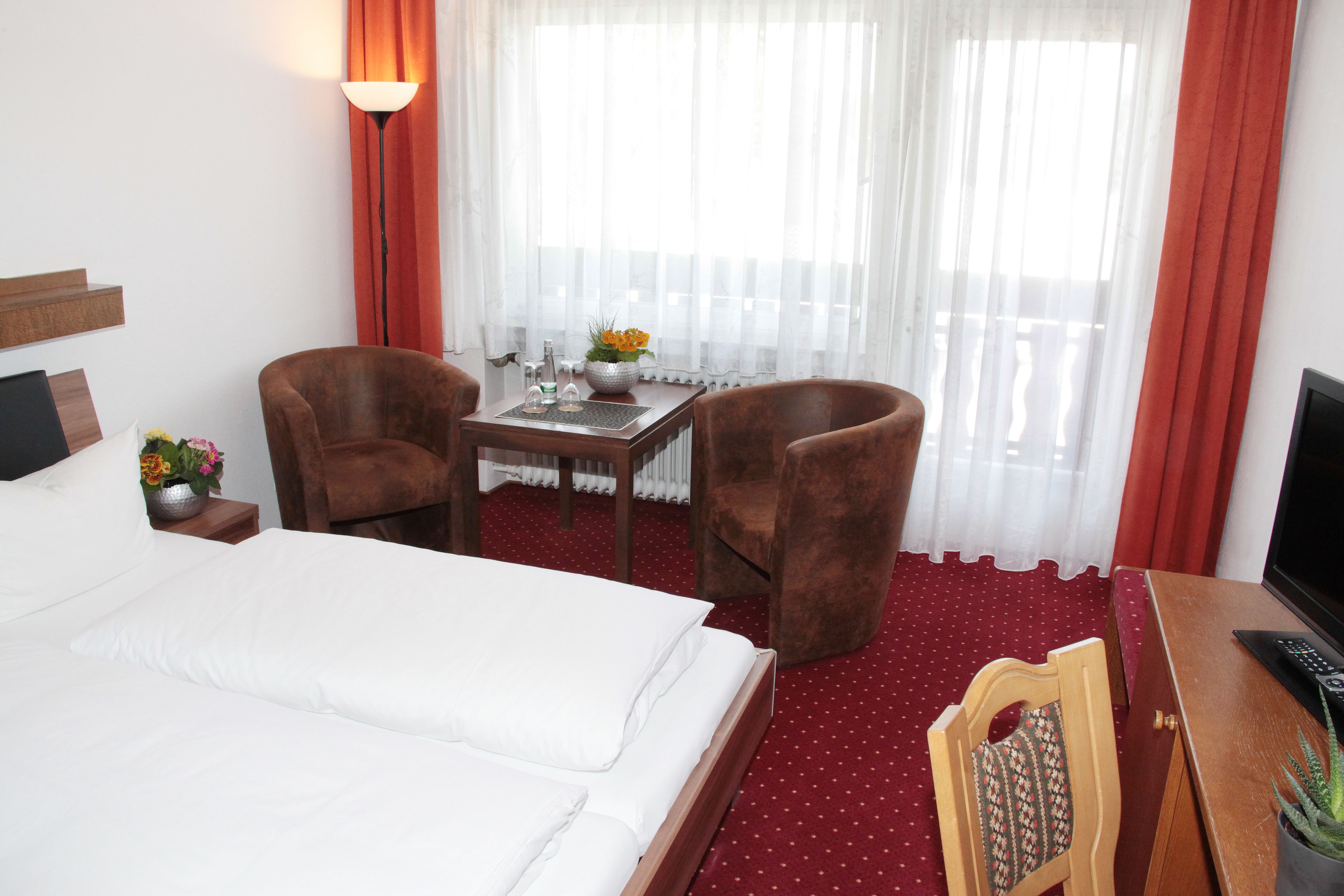 Ein Hotelzimmer mit einem Bett, zwei Sesseln, einem kleinen Tisch mit Blumenvase, einem Fernseher und einem roten Teppich.