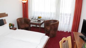Ein Hotelzimmer mit einem Bett, zwei Sesseln, einem kleinen Tisch mit Blumenvase, einem Fernseher und einem roten Teppich.