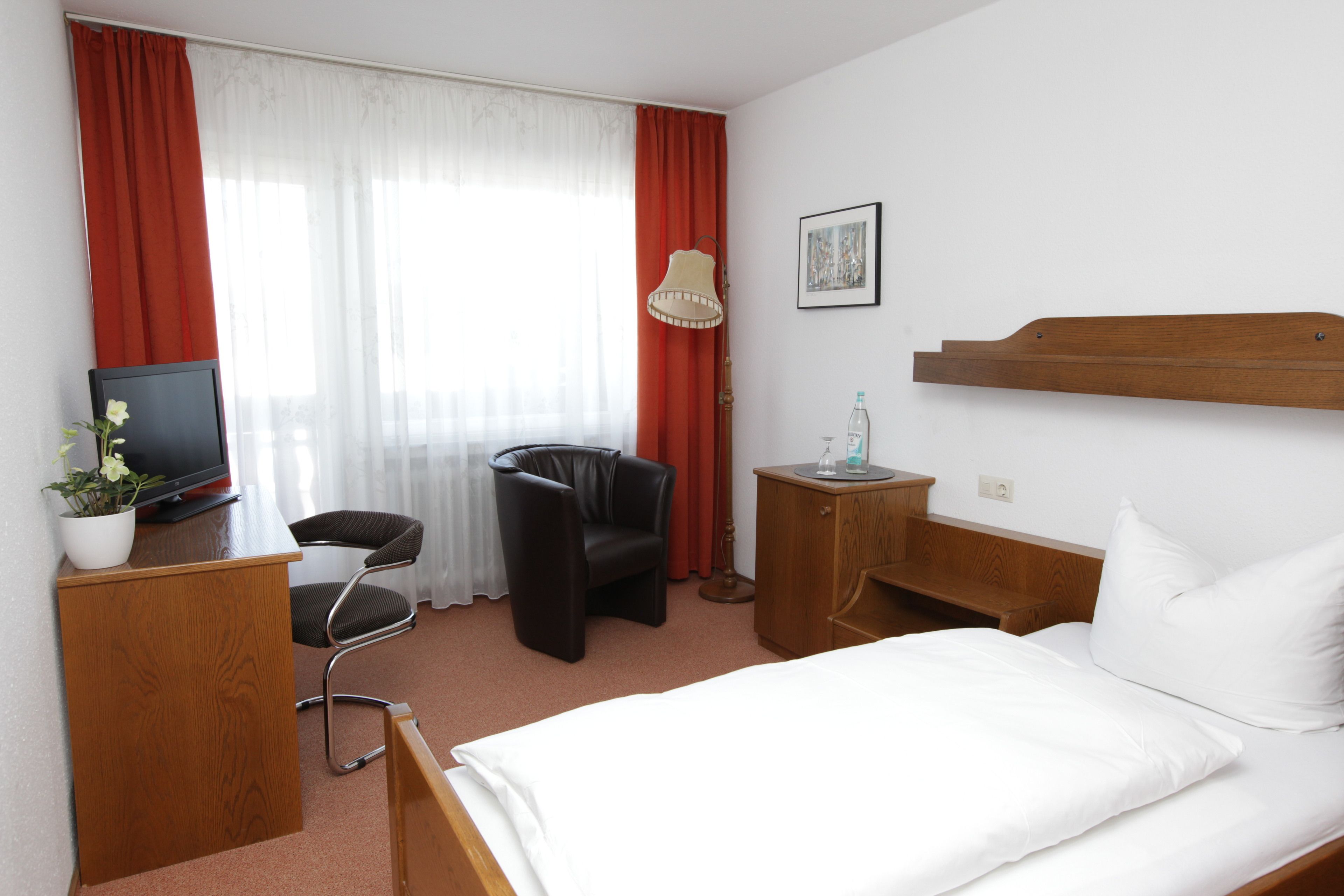 A hotel room with a bed, a desk with a computer, a chair, a lamp, and a small cabinet.