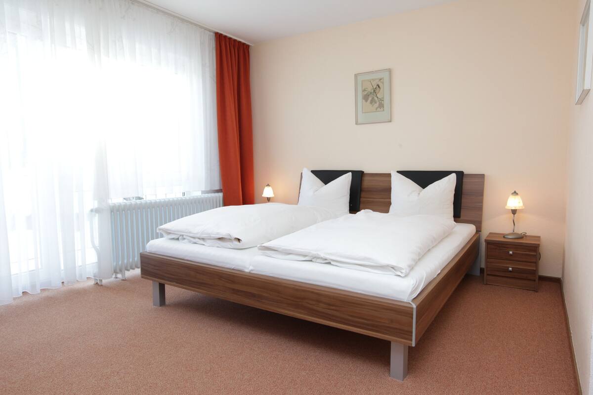 Quarto de hotel com cama de madeira, duas mesas de cabeceira com luminárias, um radiador e uma janela com cortinas.