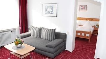 A room with a grey sofa, a small table with a flower vase, a bed with white bedding, and a red carpet.
