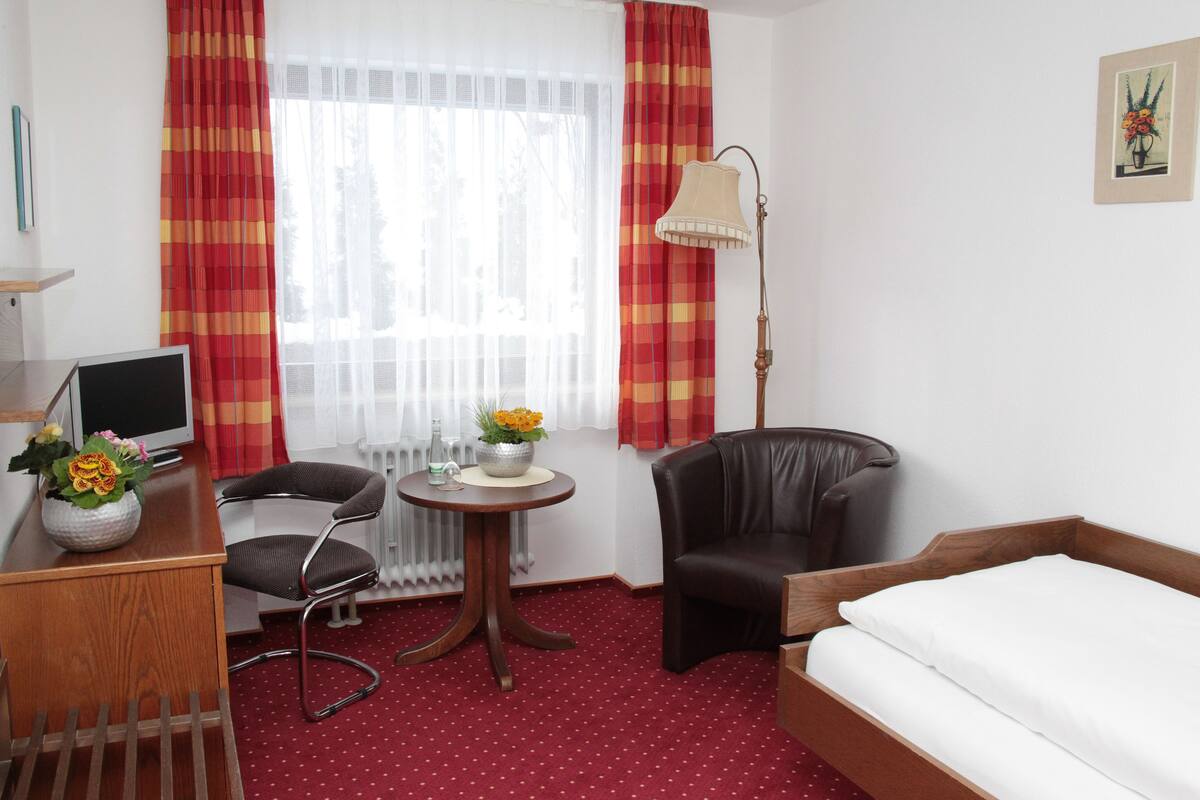 Quarto de hotel com cama, escrivaninha com computador, cadeira, mesa pequena com flores e luminária.