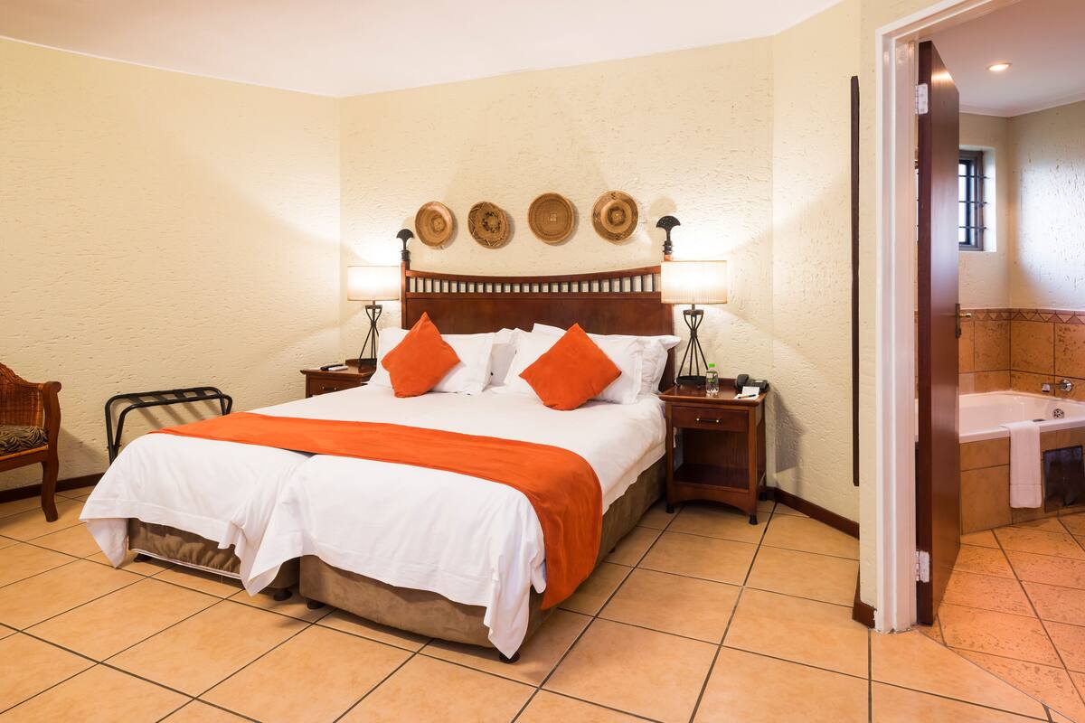 A hotel room with a large bed, two bedside tables, a nightstand, a chair, and a bathroom visible through an open door.