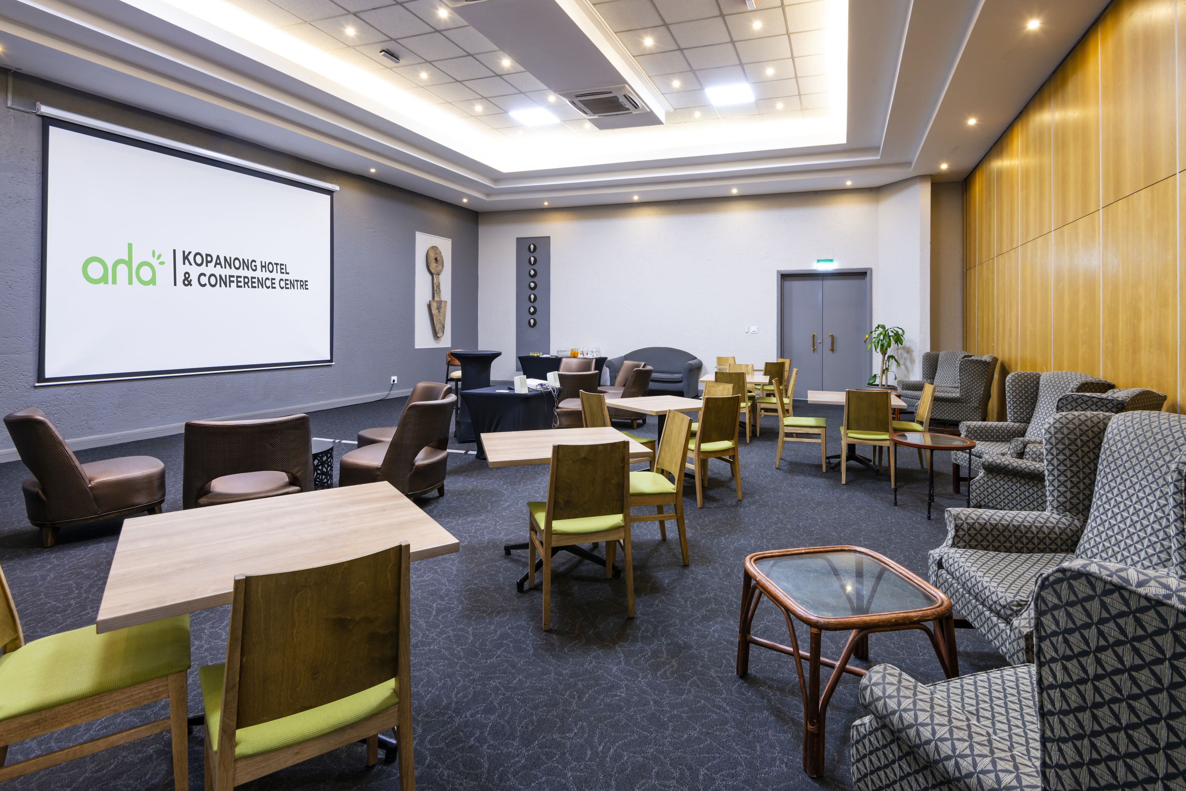 Une salle de conférence dotée d’un grand écran affichant la mention « anda | KOPANONG HOTEL & CONFERENCE CENTRE », de tables et de chaises en bois, ainsi que d’un coin salon avec un canapé à motifs.