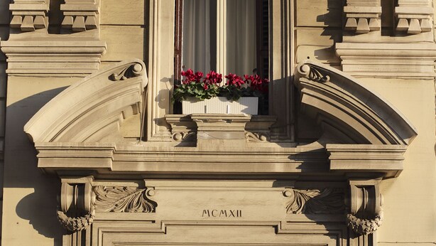 Une façade d’immeuble avec une grande porte en bois, des sculptures en pierre travaillées et une fenêtre ornée de fleurs.