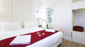 A hotel room with a neatly made bed, a red throw blanket, and a small table with a brown bag.
