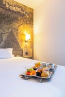 Una habitación de hotel con cama, una bandeja de desayuno, un mural en la pared y una lámpara fijada a la pared.