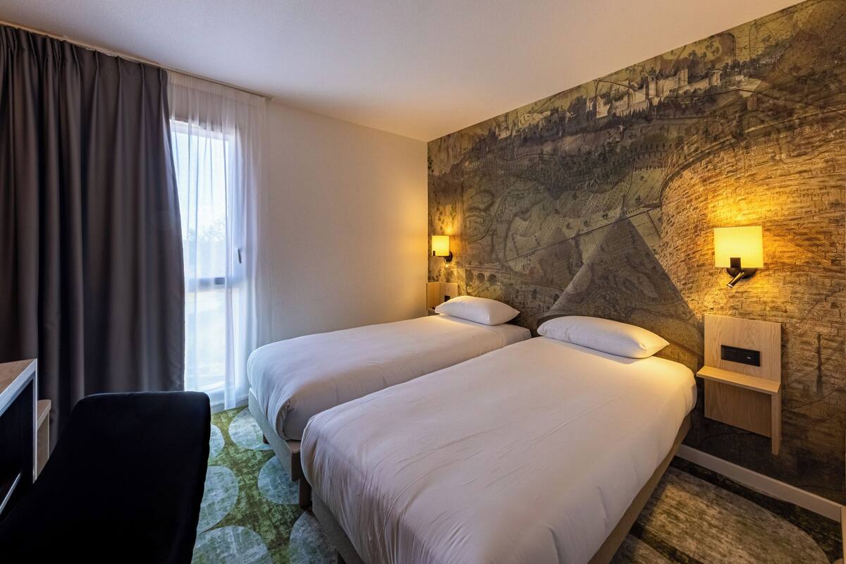 Une chambre d’hôtel avec deux lits, un bureau et une chaise. Il y a deux lampes fixées au mur et une grande fresque sur le mur.