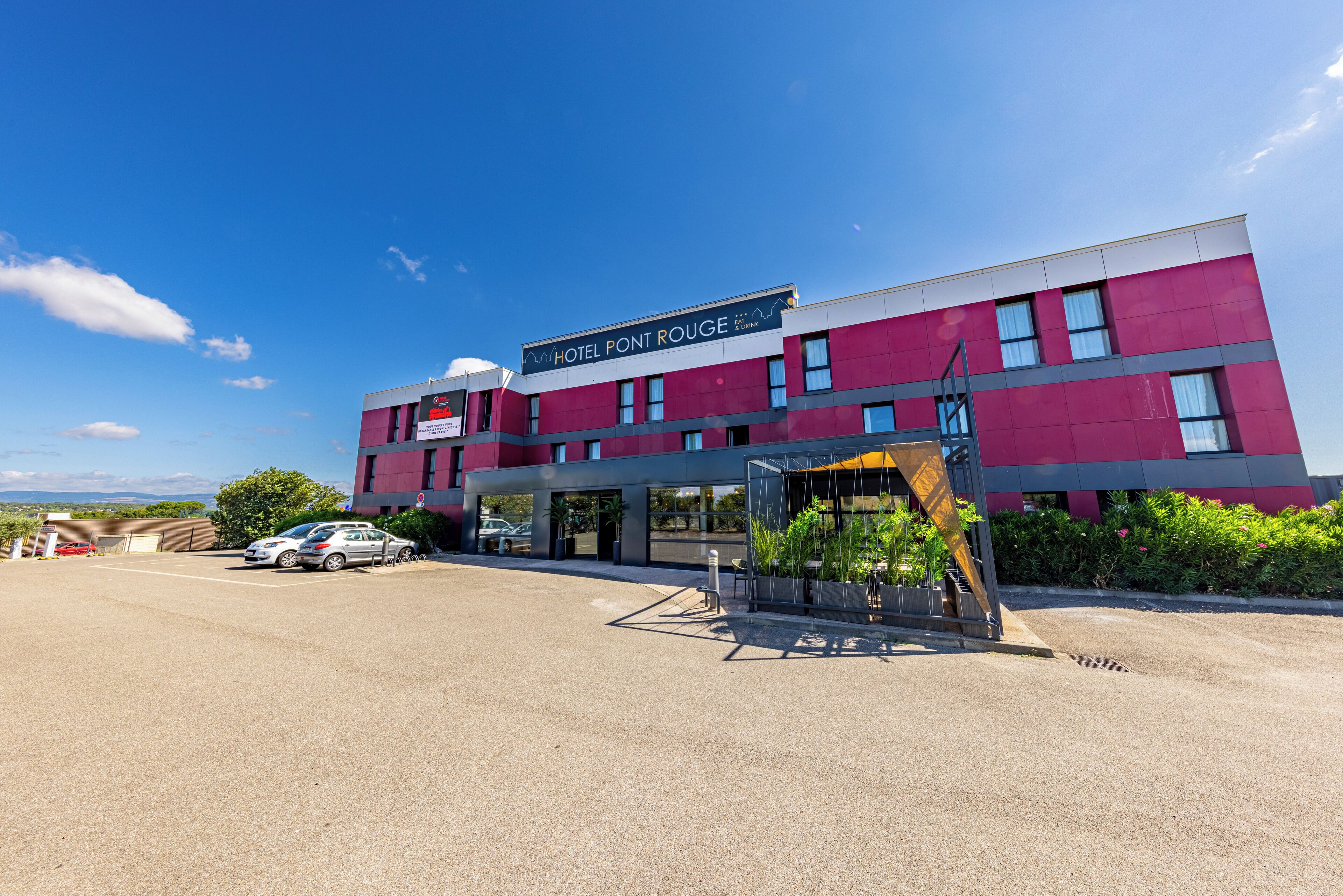 Een modern hotel met een opvallend bord waarop 'HOTEL PONT ROUGE' staat, en een parkeerplaats met verschillende auto's.
