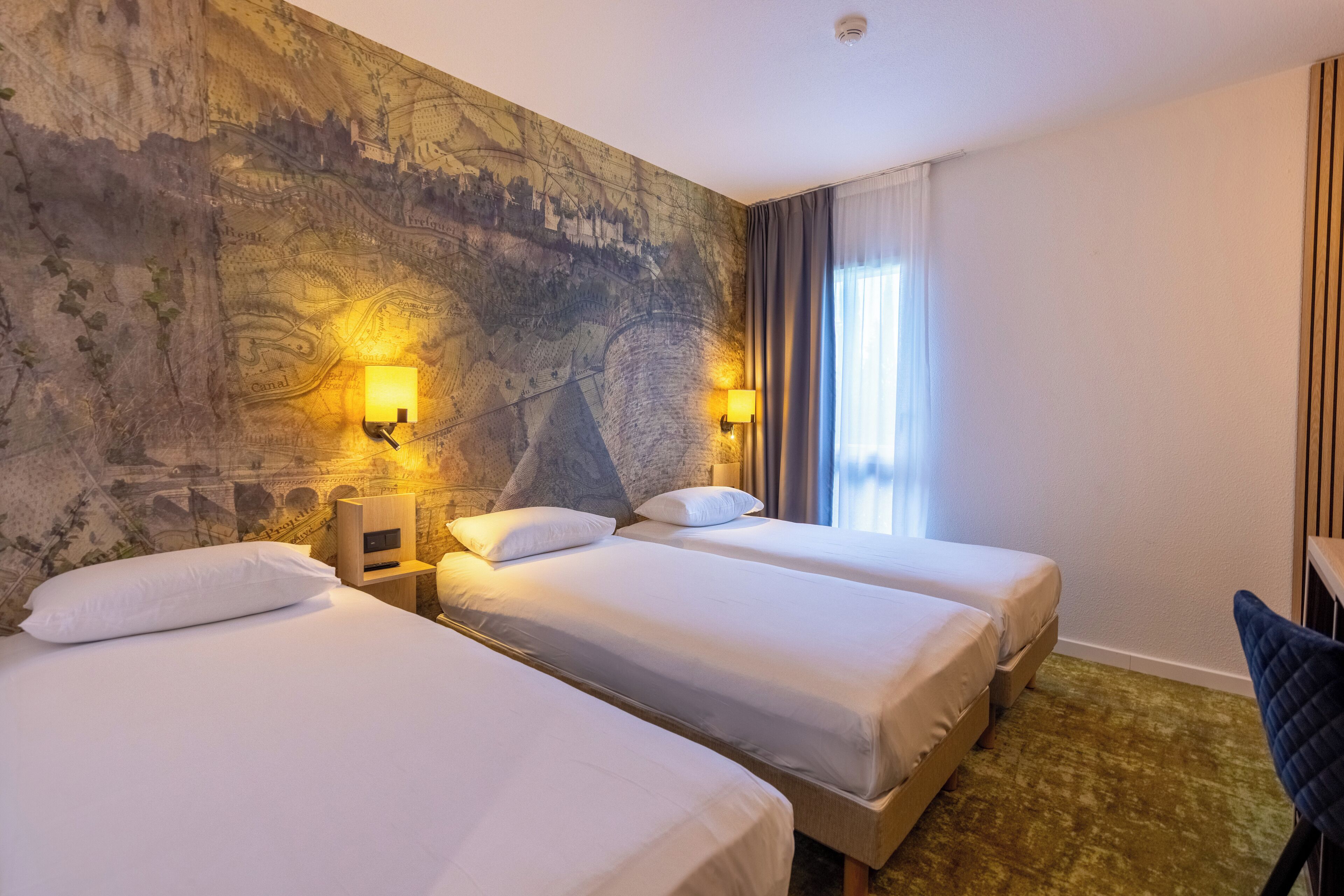 A hotel room with two single beds, a large map mural on the wall, and a desk with a chair.