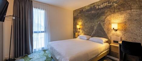 Habitación de hotel con una cama grande, un escritorio con silla, un televisor y una ventana con cortinas.