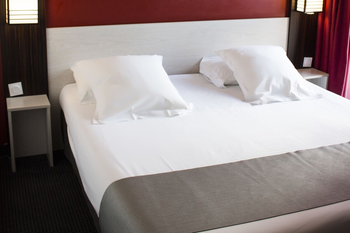 Habitación de hotel con cama, ropa de cama blanca, una pared de acento roja, un cuadro abstracto moderno y dos lámparas de noche.